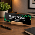 Black acrylic desk name plate with Manny Wilson text, green floral border, wood stand