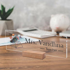 Transparent acrylic name plate, Mrs. Vandhna and Teacher text with floral book illustration.