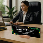 Black acrylic desk name plate with Manny Wilson and manager text, green floral border, wood stand