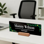 Black acrylic desk name plate with Manny Wilson manager text, green floral border, wood stand