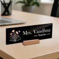 Black acrylic desk name plate: Mrs. Vandhna Teacher with floral book icon and wooden stand