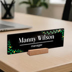 Black acrylic desk name plate on wood stand, Manny Wilson manager text, green floral border